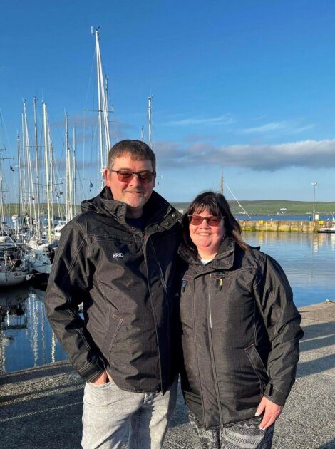 A man and woman wearing jackets stand together by a marina with several sailboats and a clear blue sky in the background.