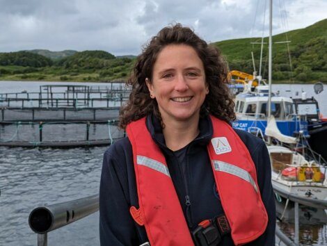 A person in a red life jacket stands near a body of water with boats and a fish farm in the background. Trees and hills are visible in the distance.