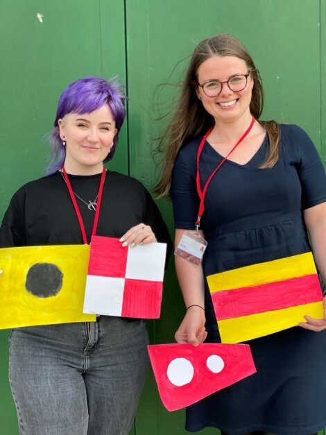 Two individuals with lanyards hold colorful, painted signal flags against a green background.