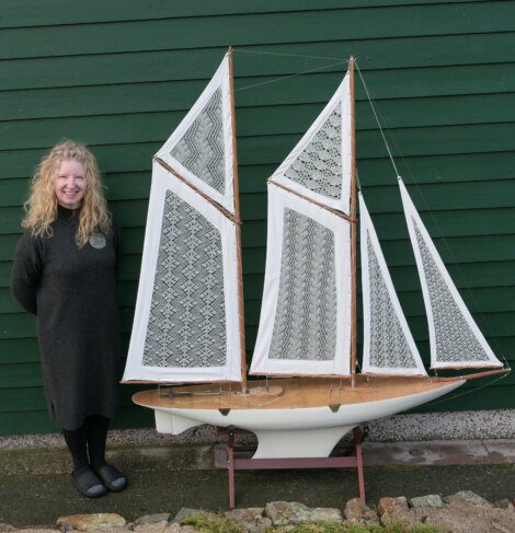 A person with curly hair stands next to a large model ship with intricate lace sails, against a green wooden wall.