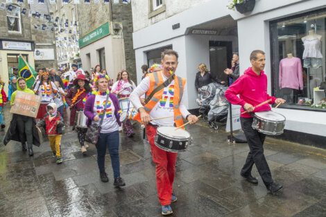 A group of people, some in colorful clothing, march on a wet street. Two men lead with drums. Flags and decorations hang above the street. Others, including children, follow in the background.