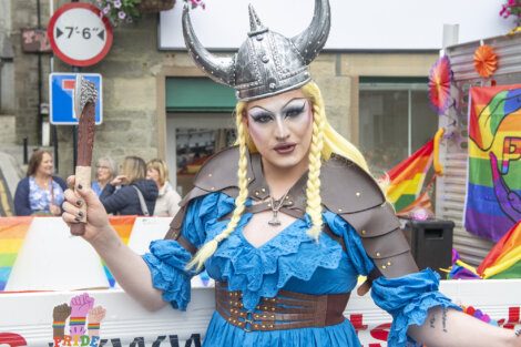 A person in a blue dress and Viking helmet, wearing makeup and holding a Viking axe, stands at a pride event with rainbow flags and people in the background.