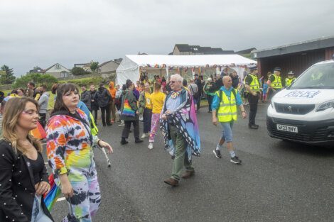 A group of people, some wearing high-visibility vests and colorful clothing, walk together near a white tent. A police vehicle and officers are visible in the background.