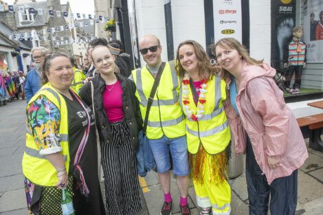 A group of five people, some in high-visibility vests, smiling and standing close together on a street with buildings and a crowd in the background.