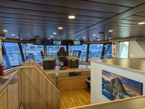 Interior view of a modern ship's bridge with navigation consoles, large windows, and mounted screens displaying various data. There is also a wooden staircase and a framed landscape picture on the wall.