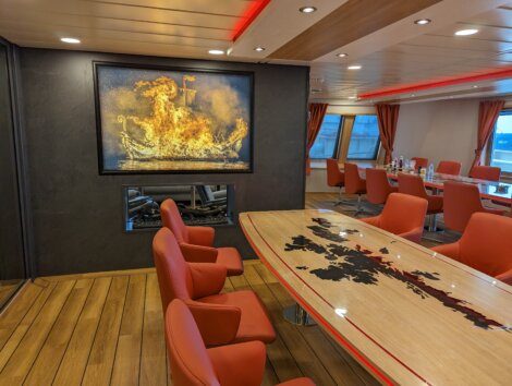 A modern conference room with red chairs, a wooden table featuring a world map, red ceiling lights, and a framed picture of a burning ship above a fireplace.