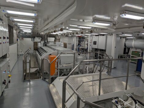 A well-lit engine room features various large machinery, control panels, and metal walkways with handrails. The equipment is neatly arranged within a clean, industrial environment.