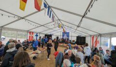 A group of people are gathering under a white tent decorated with colorful nautical flags, participating in a social event with food and drinks.