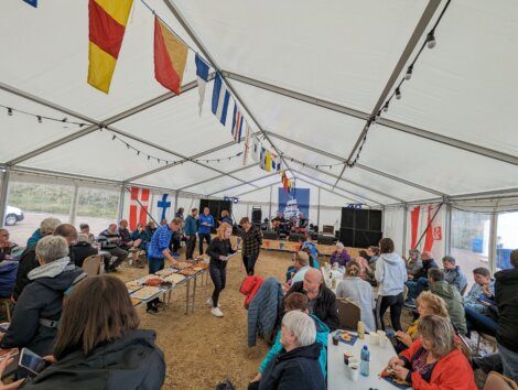 A group of people are gathering under a white tent decorated with colorful nautical flags, participating in a social event with food and drinks.