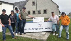 Six people stand outside a building labeled "Sub-Orbital House," holding a large check. They are dressed casually and wearing matching caps.