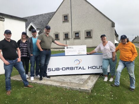 Six people stand outside a building labeled "Sub-Orbital House," holding a large check. They are dressed casually and wearing matching caps.