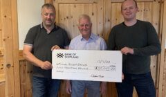 Three men standing indoors holding a large check from the Bank of Scotland made out to Nurture Room Group for fifty thousand pounds, dated 13/10/23.