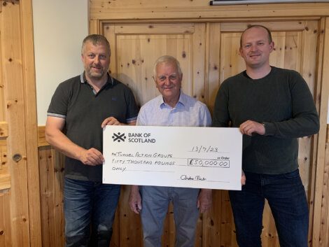 Three men standing indoors holding a large check from the Bank of Scotland made out to Nurture Room Group for fifty thousand pounds, dated 13/10/23.