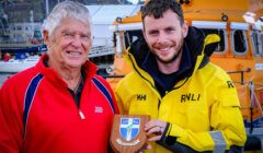 Two men stand in front of a lifeboat. The older man is in a red jacket, and the younger man, in a yellow RNLI jacket, holds a wooden plaque with a shield emblem.