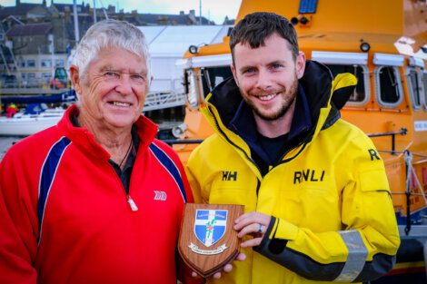 Two men stand in front of a lifeboat. The older man is in a red jacket, and the younger man, in a yellow RNLI jacket, holds a wooden plaque with a shield emblem.