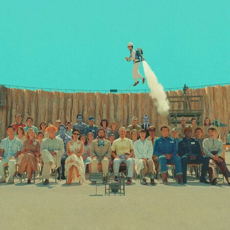 A group of people of various ages, dressed in period clothing, are seated in a row with a desert backdrop; above them, a person is flying using a jetpack.