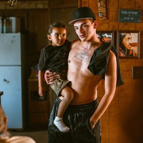 A shirtless man wearing a cap holds a young child in a kitchen with wooden walls, a visible fridge, and native-themed pictures.