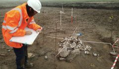 A person in an orange high-visibility jacket and white helmet is documenting an archaeological excavation site with scattered rocks and measuring tools.