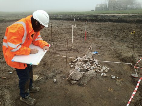 A person in an orange high-visibility jacket and white helmet is documenting an archaeological excavation site with scattered rocks and measuring tools.