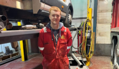 A man in a red coverall stands in a mechanic's workshop in front of a vehicle lifted for repair. Various tools and equipment are visible in the background.