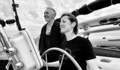 A woman and a man are steering a sailboat together. The woman is holding the wheel while the man stands beside her, both smiling. The background shows a cloudy sky and the sea.