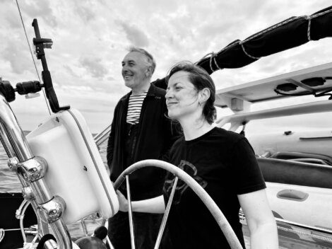 A woman and a man are steering a sailboat together. The woman is holding the wheel while the man stands beside her, both smiling. The background shows a cloudy sky and the sea.