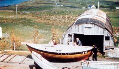 Three people work outside a curved-roof shed with boats on a hillside overlooking water and farmlands.