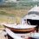 Three people work outside a curved-roof shed with boats on a hillside overlooking water and farmlands.