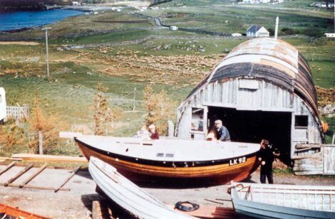 Three people work outside a curved-roof shed with boats on a hillside overlooking water and farmlands.