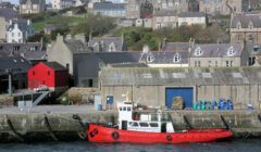 A red and white boat is docked at a waterfront in front of a row of buildings, including a prominent red warehouse, in a coastal town.