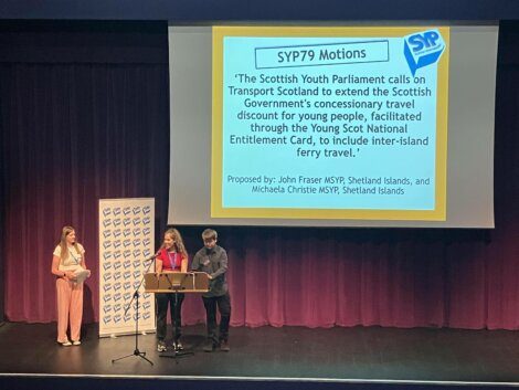 Three individuals stand on a stage with microphones. Behind them is a screen displaying a motion regarding travel discounts for young people in Scotland, presented at a Scottish Youth Parliament meeting.