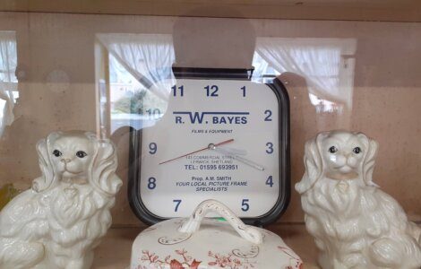 A square clock with "R.W. Bayes Films & Equipment" and a contact number is displayed between two ceramic dog figurines. The clock shows 11:12. White curtains and a street view are visible in the background.