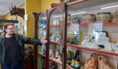 A person in a leather jacket stands beside a glass display cabinet filled with various ceramic and porcelain figurines and decorative items. The person gestures towards the objects inside.