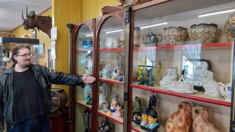 A person in a leather jacket stands beside a glass display cabinet filled with various ceramic and porcelain figurines and decorative items. The person gestures towards the objects inside.