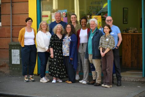 A group of 10 people stand in front of a store with green-framed windows. They are wearing casual attire and smiling at the camera.