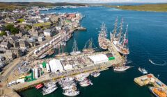 Aerial view of a bustling harbor with multiple sailing ships docked alongside a pier, surrounded by buildings and rolling hills under a partly cloudy sky.