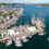 Aerial view of a bustling harbor with multiple sailing ships docked alongside a pier, surrounded by buildings and rolling hills under a partly cloudy sky.