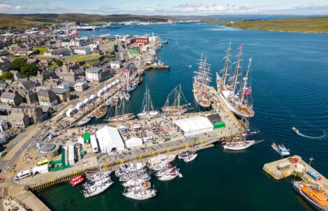 Aerial view of a bustling harbor with multiple sailing ships docked alongside a pier, surrounded by buildings and rolling hills under a partly cloudy sky.