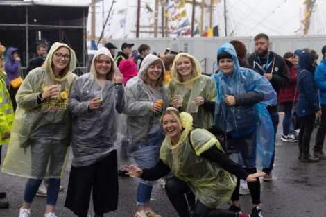 A group of people wearing colorful rain ponchos are smiling and posing for the camera at an outdoor event, amid a crowd and tall ships in the background.