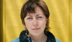 A person with short brown hair wearing a denim jacket stands in front of a bright yellow wall, looking directly at the camera.