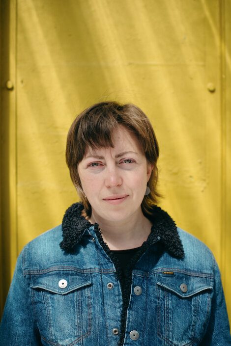 A person with short brown hair wearing a denim jacket stands in front of a bright yellow wall, looking directly at the camera.