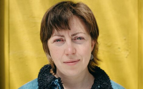 Woman with short brown hair and fair skin standing in front of a yellow background, wearing a blue jacket with a dark collar.