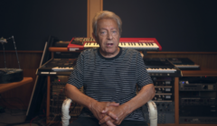 A person with gray hair sits on a chair in a music studio with multiple keyboards and audio equipment in the background. They are wearing a black and white striped shirt.