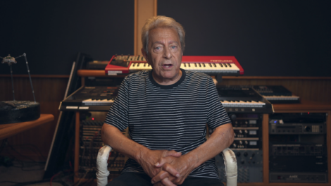 A person with gray hair sits on a chair in a music studio with multiple keyboards and audio equipment in the background. They are wearing a black and white striped shirt.