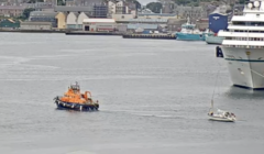 An orange tugboat guides a small sailboat near a large docked cruise ship in an industrial port area with various buildings and vessels in the background.