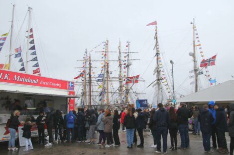 People are lined up at a catering truck on a cloudy day, with several tall ships adorned with flags in the background.