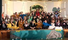 A group of people, mostly children, sit and stand on a decorated stage in costumes, likely performing a play. The backdrop includes trees and decorative elements.