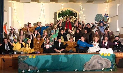 A group of people, mostly children, sit and stand on a decorated stage in costumes, likely performing a play. The backdrop includes trees and decorative elements.