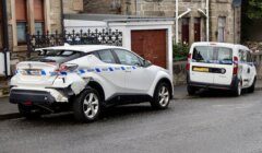 A white car with rear-end damage and a police tape around it is parked on a street next to a white van.