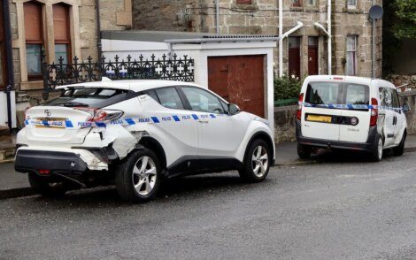 A white car with rear-end damage and a police tape around it is parked on a street next to a white van.